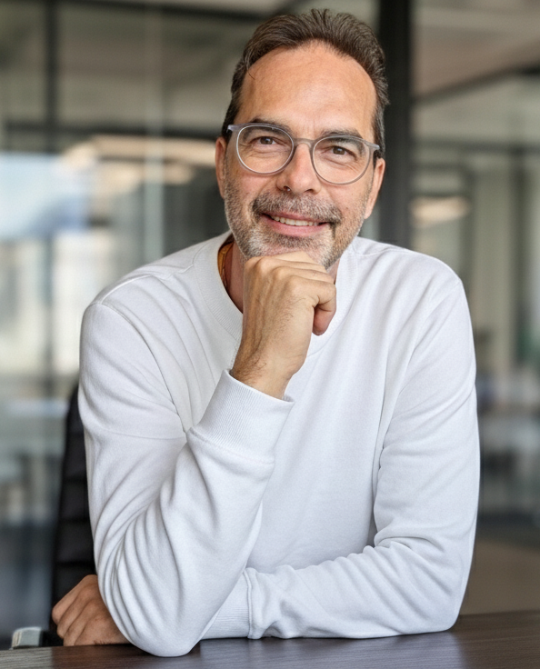 Freundlicher Psychotherapeut mittleren Alters mit Brille und weißem Pullover sitzt lächelnd an einem Schreibtisch in einer modernen Praxis in Remscheid.