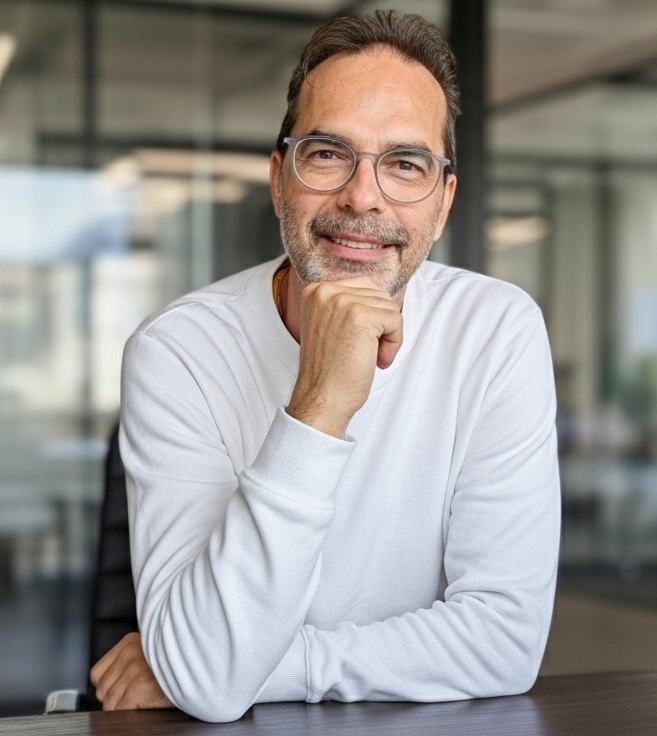 Freundlicher Psychotherapeut mittleren Alters mit Brille und weißem Pullover sitzt lächelnd an einem Schreibtisch in einer modernen Praxis in Remscheid.